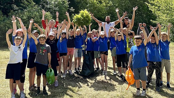 Kinder und Erwachsene um einen frisch gepflanzten Baum rechten die Arme in die Höhe
