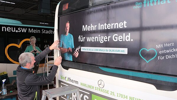 Ein Mann auf einer Leiter beklebt einen Bus mit Werbung für fitfalt. 