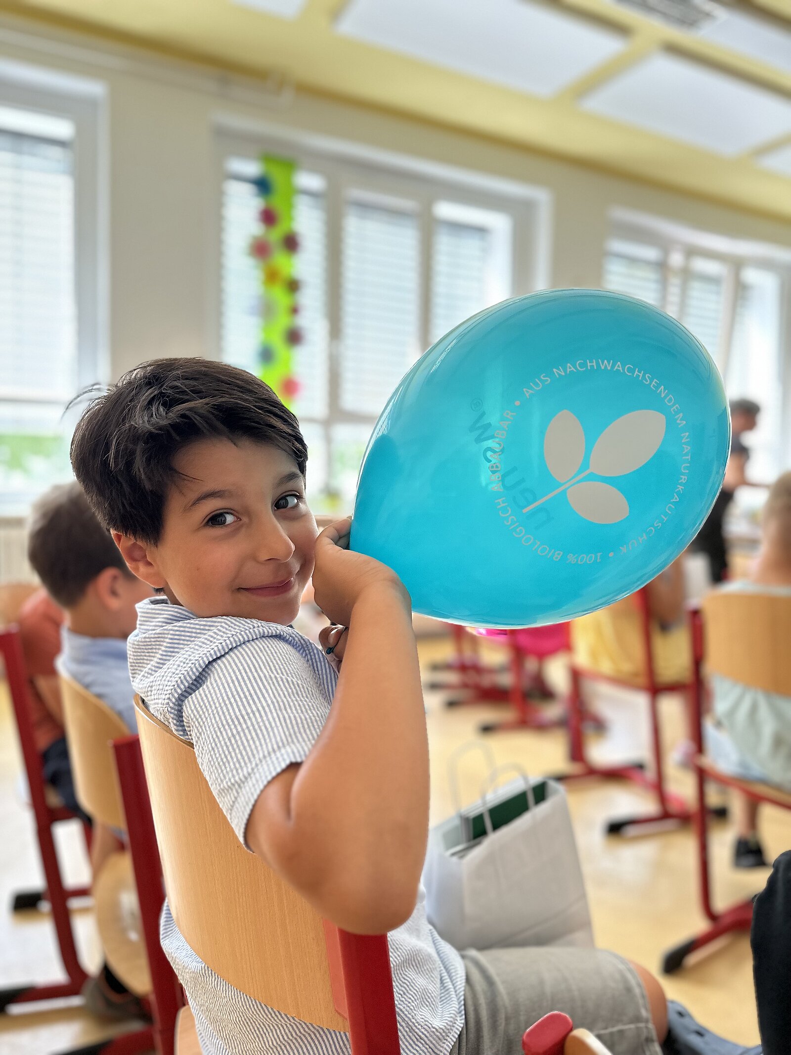 lächelnder Junge in einem Klassenraum hält einen türkis-farbenen neu.sw Luftballon in der Hand