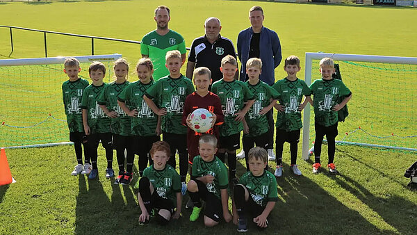 Gruppenfoto einer Kinderfußballmannschaft des SV Neuenkirchen mit drei Trainern und Betreuern. Die Jungen und Mädchen tragen grüne Trikots. Der Torwart in der Mitte trägt dunkelrot und hält einen Fußball in der Hand. 