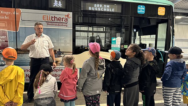 Mädchen und Jungen hören Erklärungen des Busschullehrers, der vor ihnen steht.  Dahinter ein Linienbus.