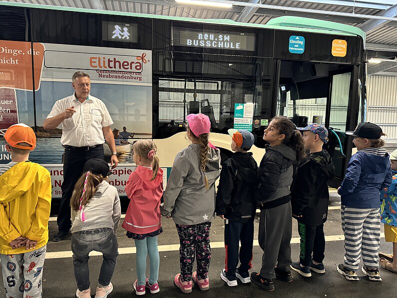 Mädchen und Jungen hören Erklärungen des Busschullehrers, der vor ihnen steht.  Dahinter ein Linienbus.