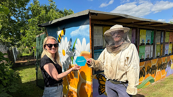 Vor einem bunten Bienenwagen übergibt eine junge Frau eine Papier-Bienenwabe mit neu.sw Logo an den Imker. 