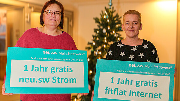 Zwei Frauen mit einem Gewinn-Scheck von neu.sw vor einem Weihnachtsbaum