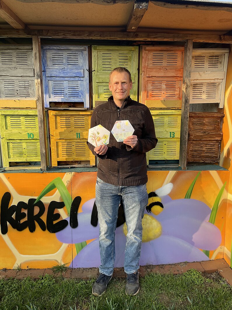 Imker Stefan Potraz steht vor den bunt bemalten Bienenstöcken und hält die beiden Bienenwaben in die Kamera, die aus der neu.sw Patenklasse stammen. 