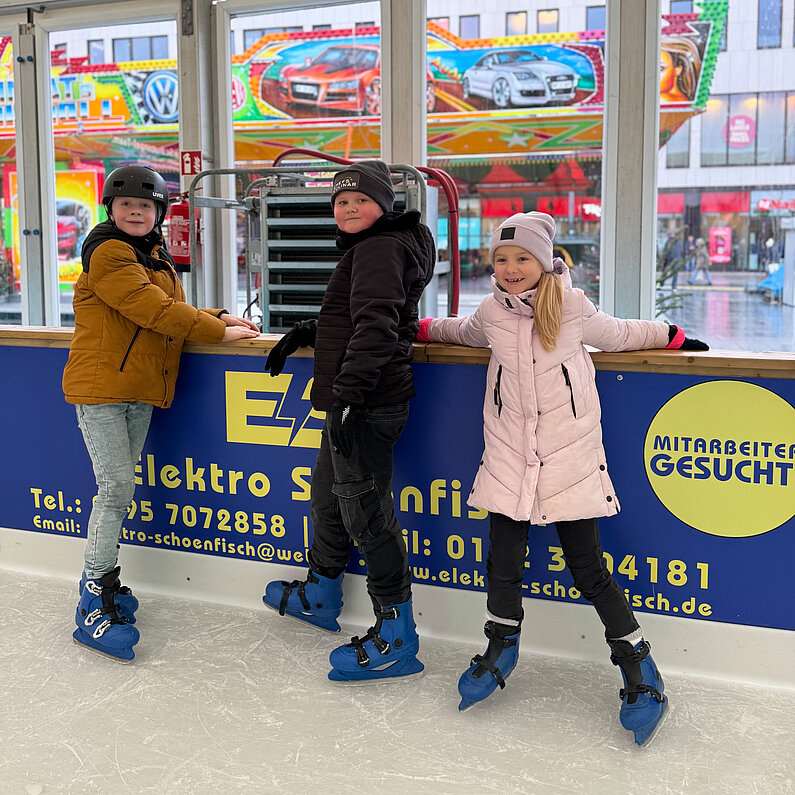 Drei Kinder ruhen sich an der Bande der Eislauffläche aus. 