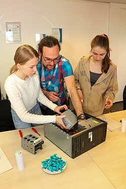 Mann mit Brille und Dreadlocks erklärt zwei jungen Frauen das Computer-Innenleben. .