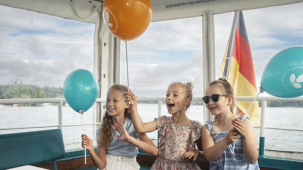 Drei Kinder mit blauen und orangen Luftballons im neu.sw Linienschiff.