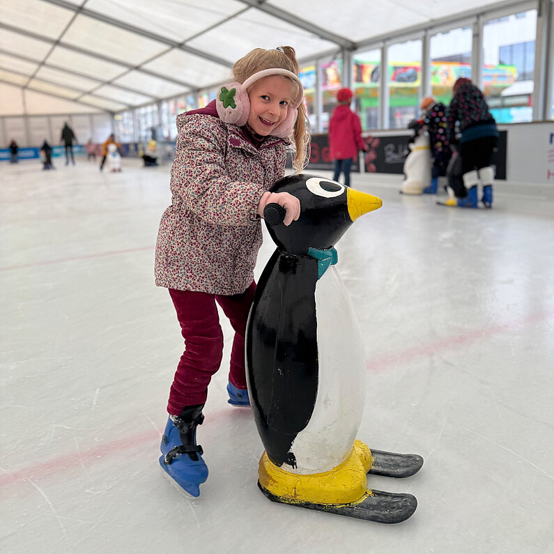 Ein Mädchen fährt Schlittschuh und hält dabei eine Eislaufhilfe in Form eines Pinguins.