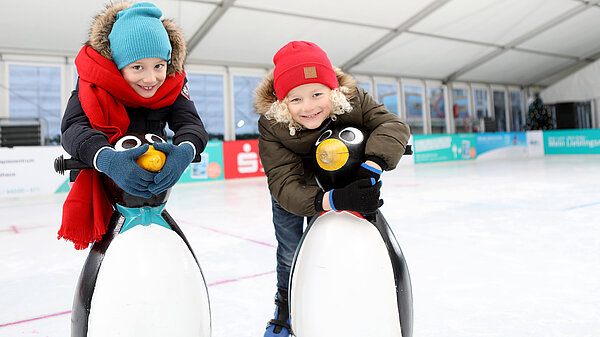 Zwei lächelnde Kinder in Wintersachen mit Lauflernhilfen in der neu.sw Eislaufhalle.
