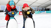 Zwei lächelnde Kinder in Wintersachen mit Lauflernhilfen in der neu.sw Eislaufhalle.