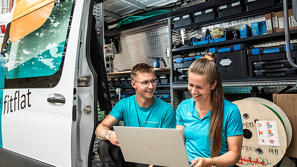 Mitarbeiterin und Mitarbeiter in türkis-farbenen neu.sw Shirts sitzen mit Laptop in einem neu.sw Technik-Transporter.