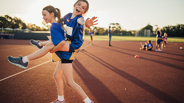 Ein Mädchen hat ein anderes Huckepack - beide in Sportkleidung im Leichtatlethikstadion