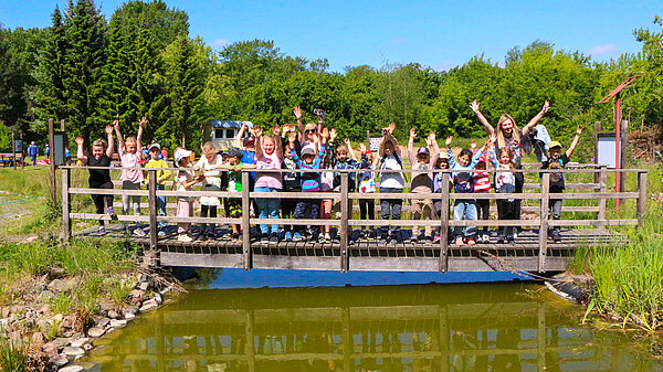 Schulklasse auf Brücke im Gruppenbild mit nach oben ausgestreckten Armen.