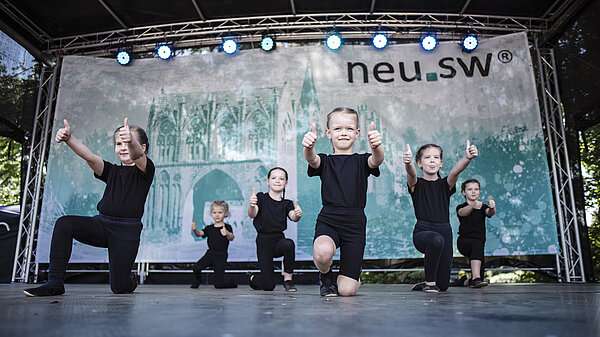 Kinder tanzend auf Bühne mit Stadtsilhouette und neu.sw Logo.