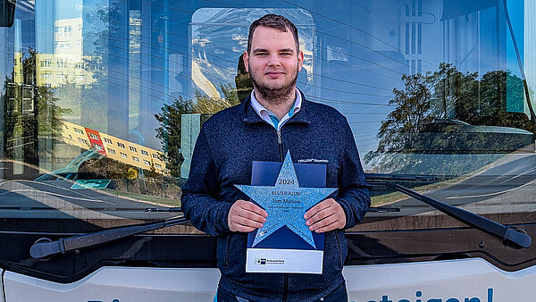 Ausgezeichneter Tom Marlow von der NVB vor einem Stadtbus mit Urkunde.