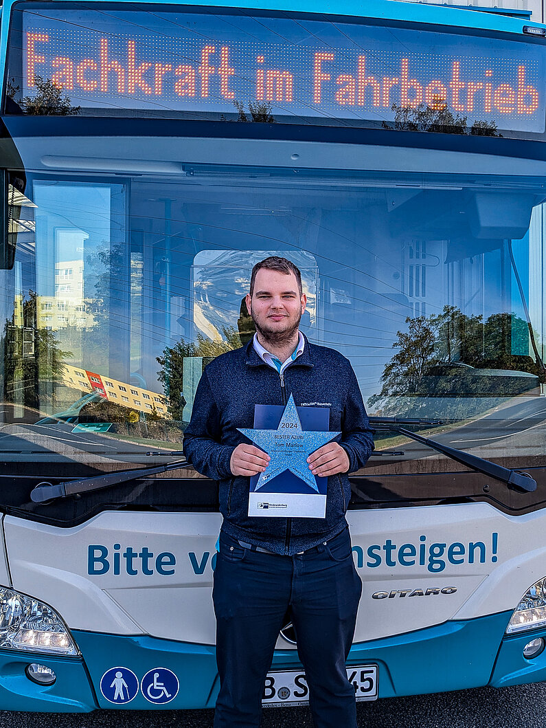 Busfahrer  vor einem Stadtbus mit der Anzeige "Fachkraft im Fahrbetrieb.