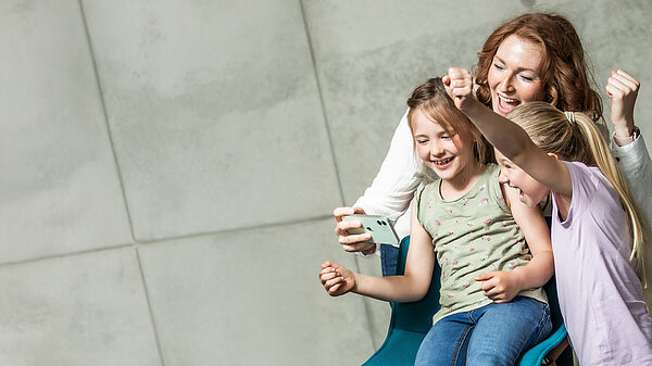 zwei Kinder und eine Frau blicken auf ein Handy und freuen sich