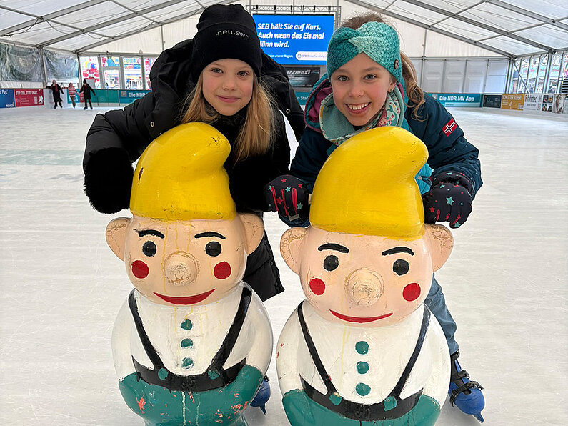 Zwei Mädchen mit Zwergen-Eislaufhilfen auf dem Eis.