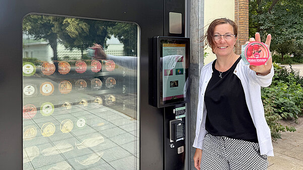 Frau lehnt am schwarzen Eisautomat und hält lächelnd einen Becher Himbeer-Schokolade-Eis in die Kamera. Durch eine Scheibe im Automaten sind die verschiedenen Eissorten in Fächern zu sehen.