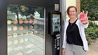 Frau lehnt am schwarzen Eisautomat und hält lächelnd einen Becher Himbeer-Schokolade-Eis in die Kamera. Durch eine Scheibe im Automaten sind die verschiedenen Eissorten in Fächern zu sehen.