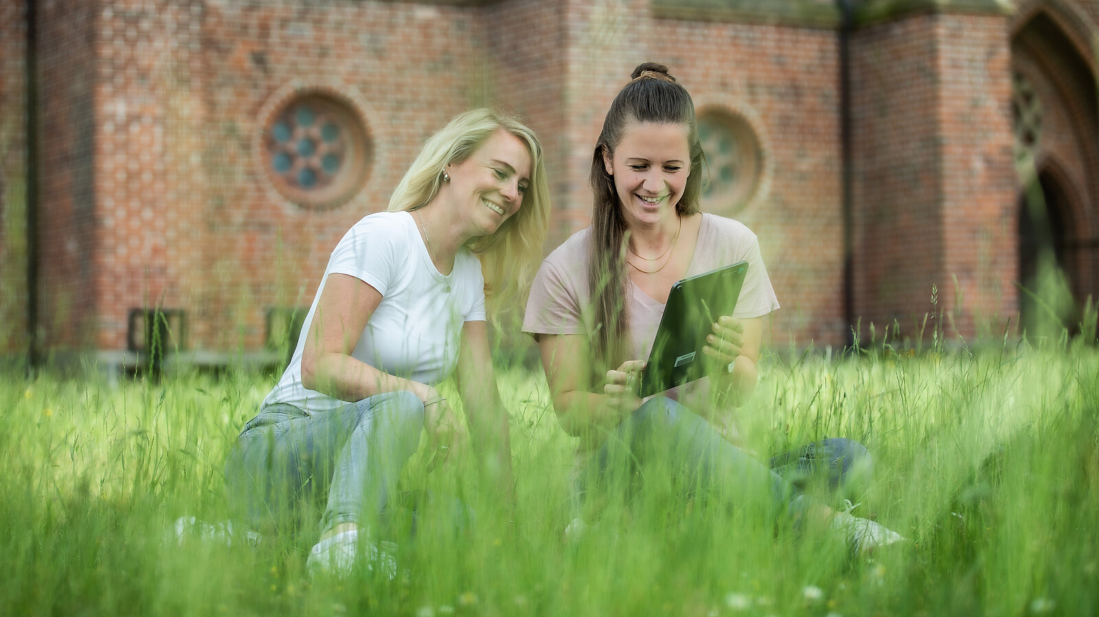 Zwei junge Frauen sitzen lachend im Gras vor einem Backsteingebäude und schauen gemeinsam auf ein Tablet.