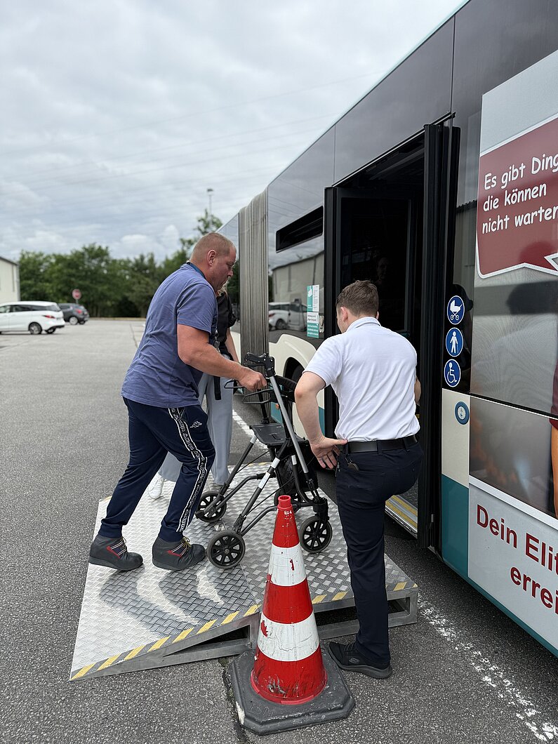 Mann mit Rollator steigt über Rampe in einen Stadtbus ein. Busfahrer assistiert. 