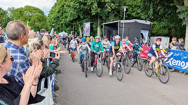 Kinder beim Radfahren in der Gruppe. Applaudierende Erwachsene an der Strecke.