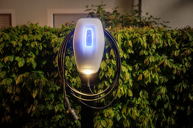 Mennekes-Werbefoto: Silberfarbene Wallbox mit blau leuchtendem Display vor einer großen Hecke. 