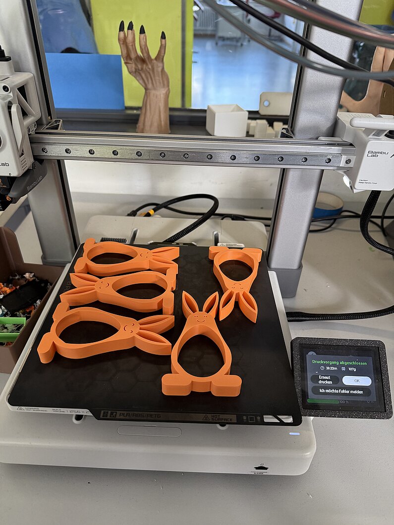 fertige orangefarbene Osterhasen auf einer Unterlage vor dem 3D-Drucker.