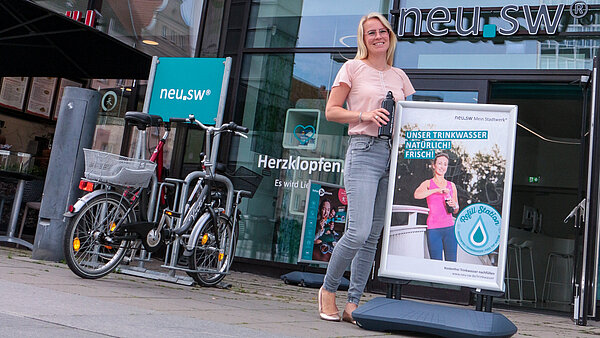 Eine junge Frau steht mit einer Mehrwegflasche an einem Plakat-Aufsteller mit "Refill"-Aufkleber vor dem neu.sw Kundenbüro. 