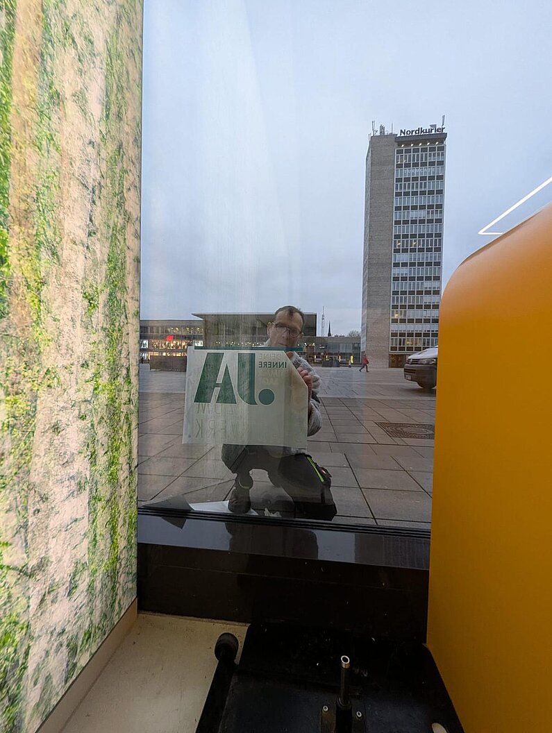 Blick von innen, aus dem Schaufenster heraus: Beauftragter Unternehmer bringt Beklebung unten in der Ecke des Schaufensters an.