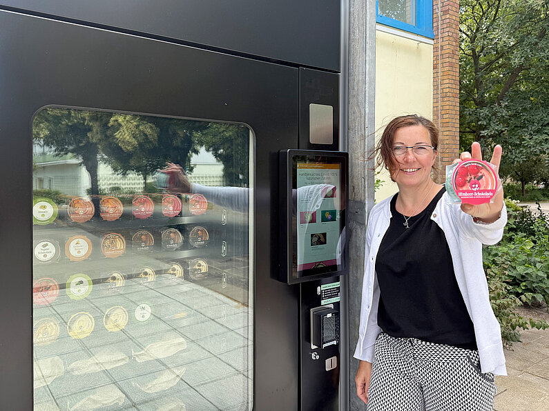 Frau lehnt am schwarzen Eisautomat und hält lächelnd einen Becher Himbeer-Schokolade-Eis in die Kamera. Durch eine Scheibe im Automaten sind die verschiedenen Eissorten in Fächern zu sehen.
