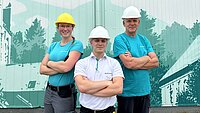 Gruppenbild: Eine Mitarbeiterin und ein Mitarbeiter in neu.sw Shirts lächenld mit verschränkten Armen vor einem neu.sw Wandbild.