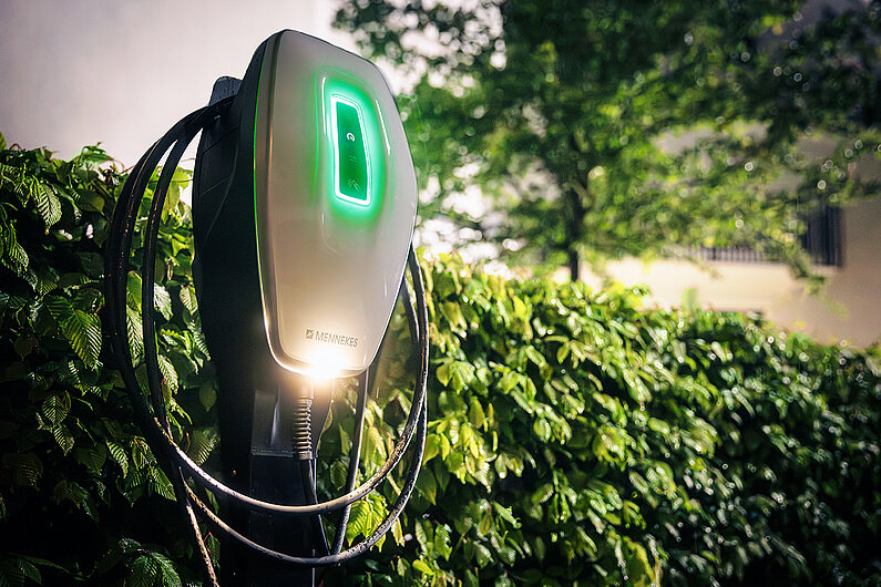 Mennekes-Werbefoto: Silberfarbene Wallbox mit grün leuchtendem Display vor einer großen Hecke. 