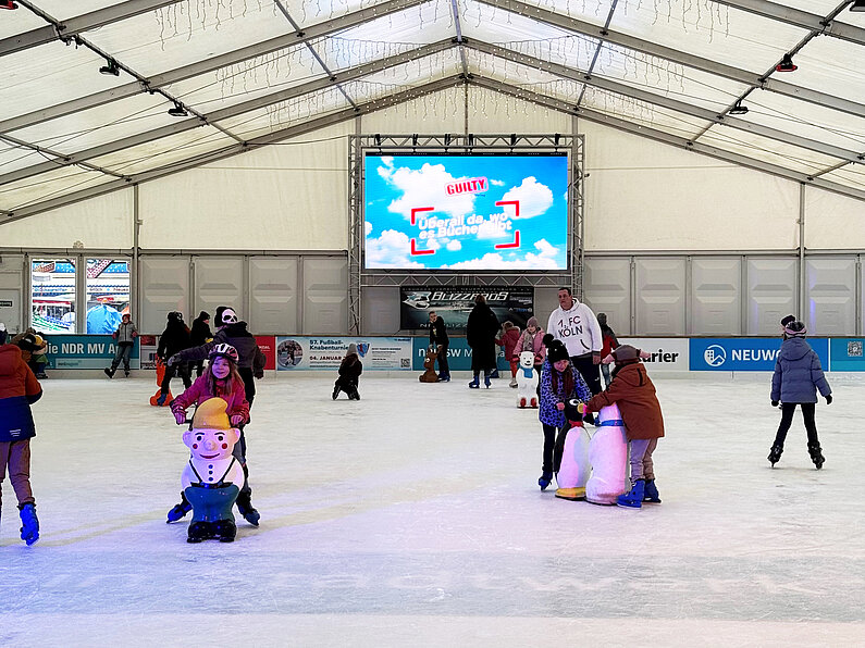 Kinder laufen Schlittschuh in der neu.sw Eislaufhalle. Einige halten Eislaufhilfen in verschiedenen Formen vor sich.