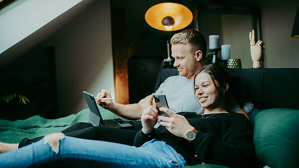 ein Mann und eine Frau sitzen auf einem Bett und halten elektronische Geräte in der Hand