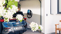 Mennekes-Werfefoto: Blick auf eine offene Garage mit dunkelblauem E-Auto Seat Cupra und dunkelblaer Ladestation an der Wand. 