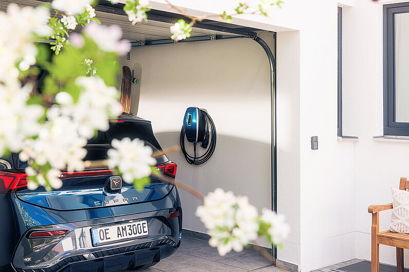 Mennekes-Werfefoto: Blick auf eine offene Garage mit dunkelblauem E-Auto Seat Cupra und dunkelblaer Ladestation an der Wand. 
