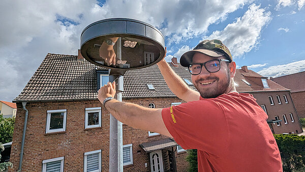 Mitarbeiter mit rotem Shirt und schwarzem Basecap mit der Auschrift "Elektrotechnik Draewe" lächelt in die Kamera, während er an der Leuchte einer Straßenlaterne schraubt. 