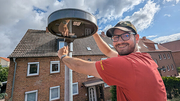 Mitarbeiter mit rotem Shirt und schwarzem Basecap mit der Auschrift "Elektrotechnik Draewe" lächelt in die Kamera, während er an der Leuchte einer Straßenlaterne schraubt. 