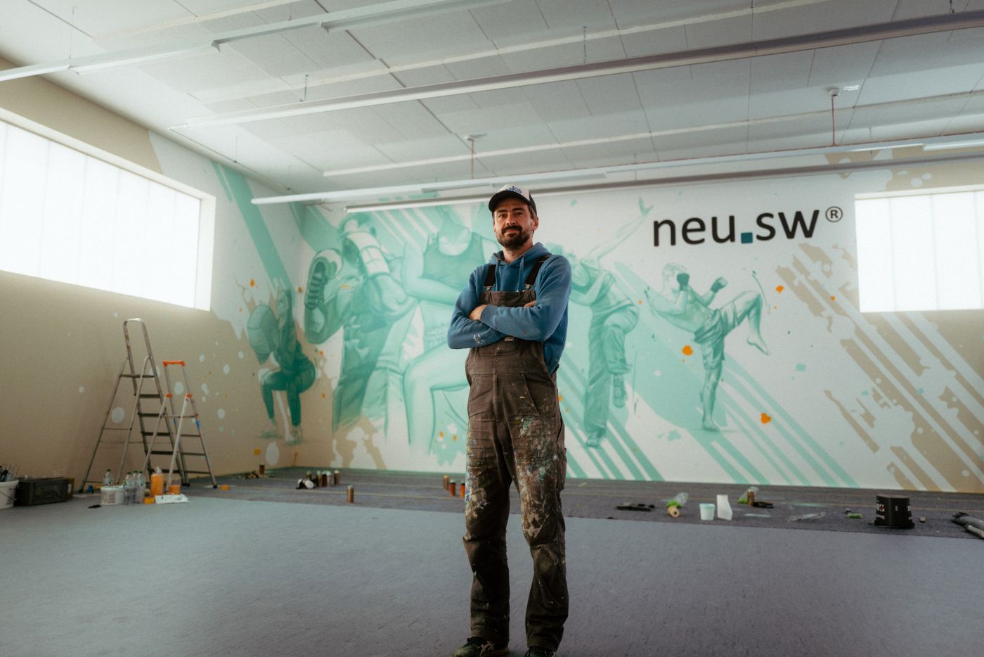 Künstler Enrico Pense steht vor seinem Werk, einem türkisfarbenen Wandbild mit verschiedenen Sportlern in Aktion.