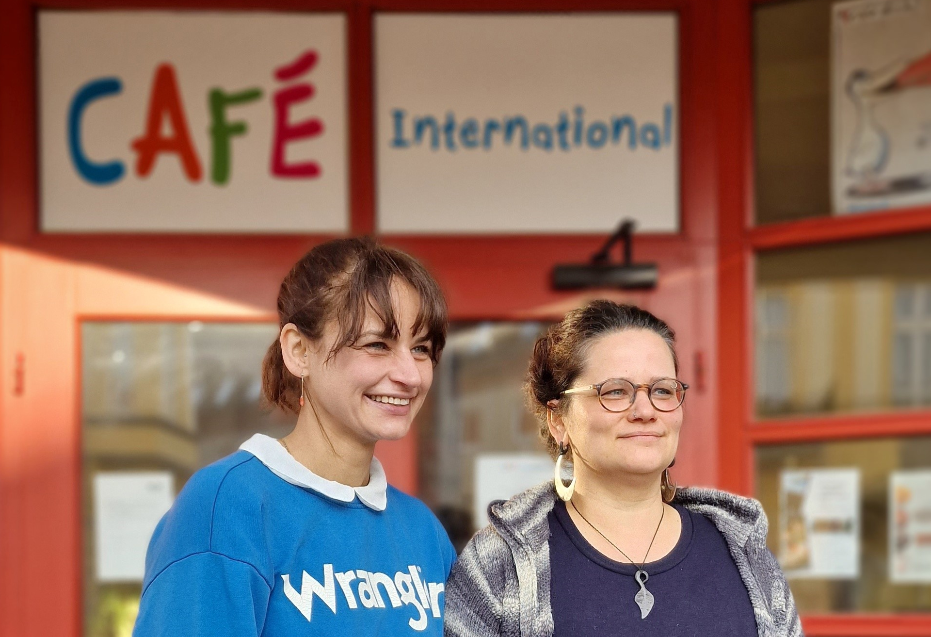 Teamleiterin Marie Ortmann und Migrationsberaterin Michelle Ruthenberg stehen lächelnd vor dem Eingang des Cafés  mit dem bunten Schriftzug "Café International"